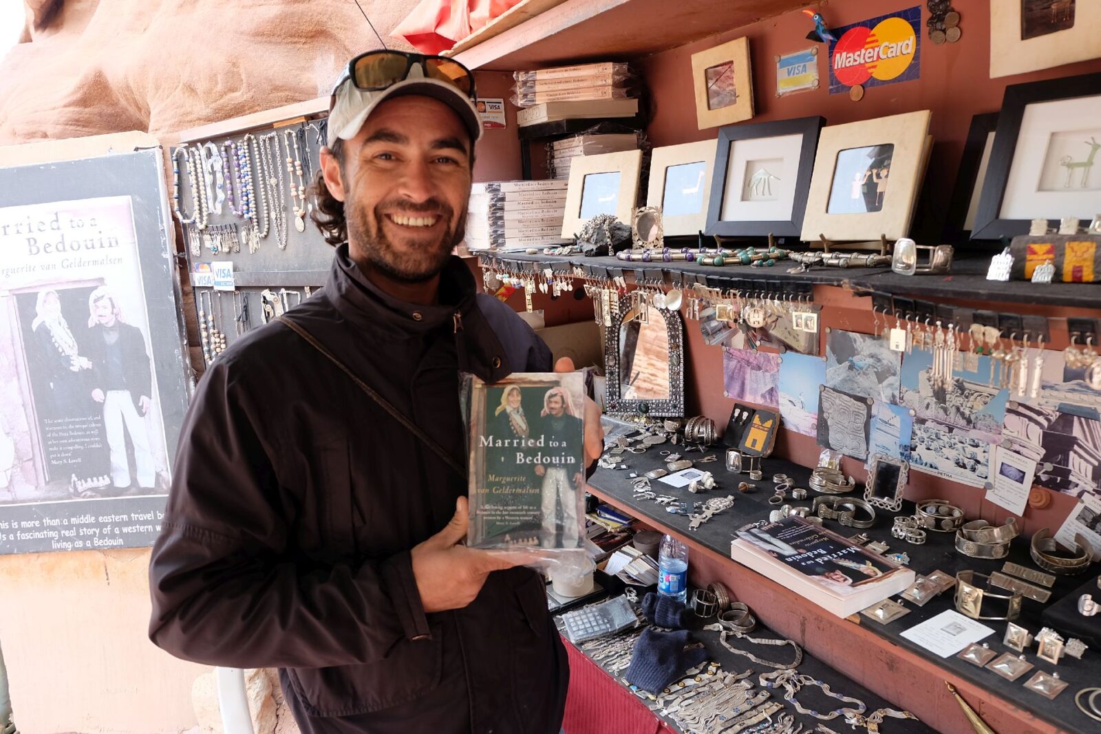 A Bedouin shopkeeper in Petra holding the book Married to a Bedouin, written by his mother, with handmade jewellery and souvenirs displayed inside his small stall.