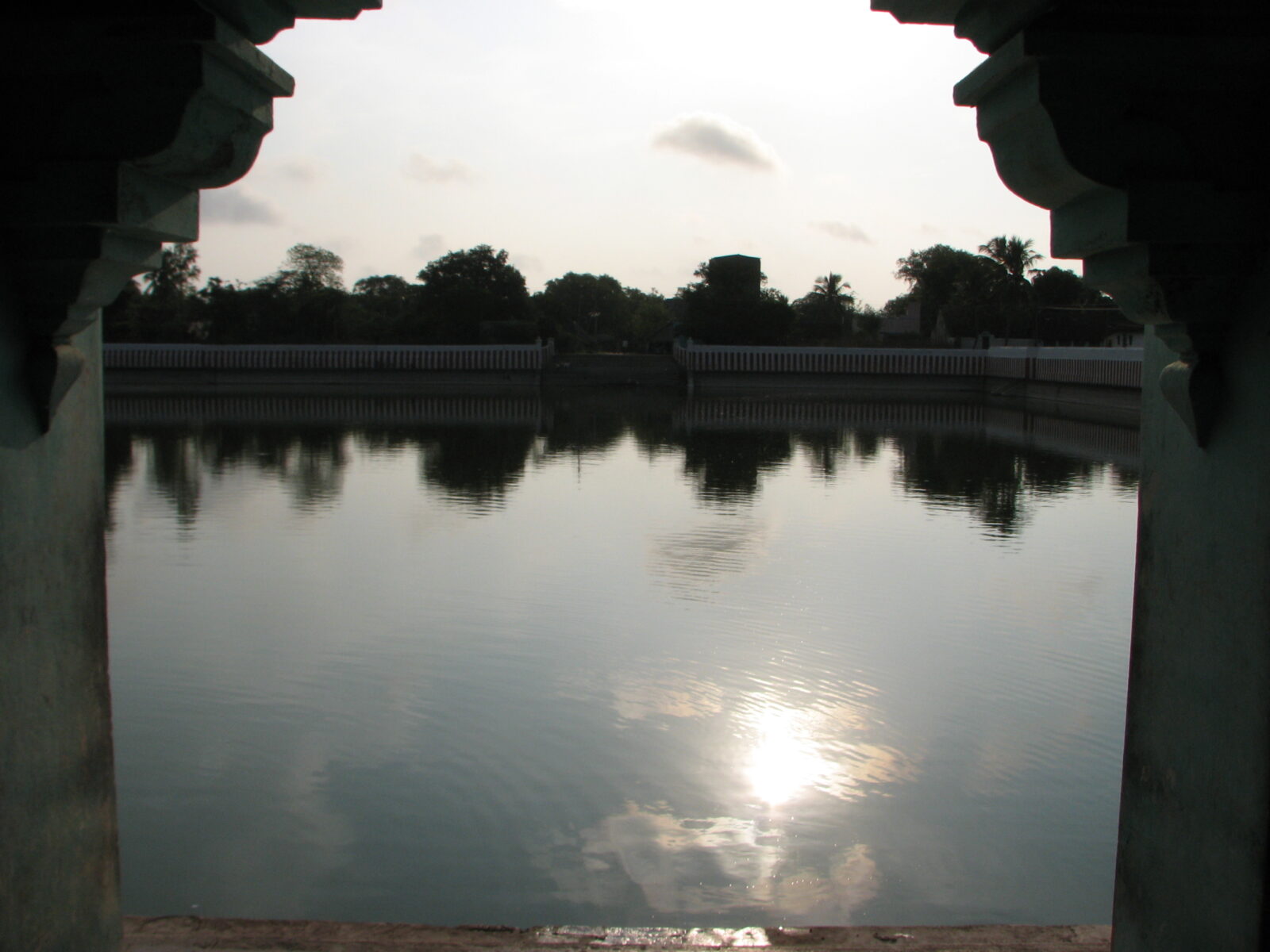 Temple tank seen through stone mandapa pillars at sunset in Tamil Nadu